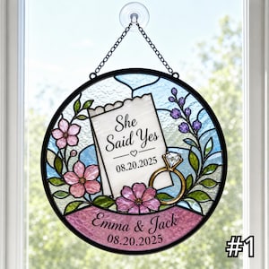 May include: A circular stained glass suncatcher featuring a white note with the text She Said Yes and 08.20.2025, a gold diamond ring, pink flowers, and purple sprigs. A pink banner at the bottom displays the names Emma & Jack and 08.20.2025.
