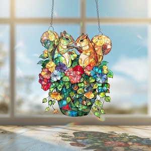 Puede incluir: Adorno de ventana de vidriera con dos ardillas entre flores coloridas en un jarrón de mosaico. Las ardillas son de color verde y naranja. Las flores son rojas, azules, moradas y amarillas. Cuelga de dos cadenas.