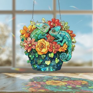 May include: A stained glass suncatcher featuring two chameleons nestled among colorful flowers. The chameleons are green and teal, surrounded by orange, red, and yellow blooms. The basket is a mosaic of blue and green glass, hanging from a chain.