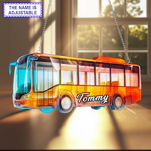 May include: A translucent orange and blue bus ornament hanging from two silver chains. The side of the bus features the name Tommy in black script lettering. A rectangular sign above the bus displays the text THE NAME IS ADJUSTABLE.