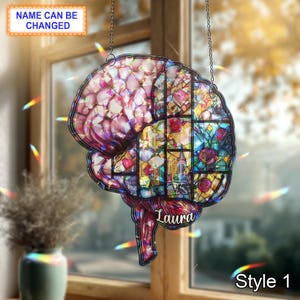 Puede incluir: Un atrapasoles con forma de cerebro de vidriera con una cadena para colgar. El diseño presenta un mosaico colorido de tonos rosas, morados, azules y verdes, con el nombre "Laura" y el texto "Style 1" visibles.