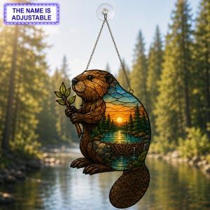 May include: A stained glass suncatcher shaped like a beaver holding a branch, featuring a sunset landscape with a river and dam inside its body. The piece hangs from a metal chain with a suction cup. Text at the top reads THE NAME IS ADJUSTABLE.