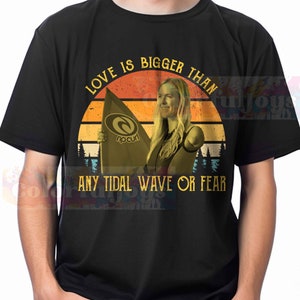 May include: A black t-shirt with a vintage-style graphic of a woman surfing. The graphic includes a sunset, a surfboard with the text "rip curl", and the text "Love is bigger than any tidal wave or fear".