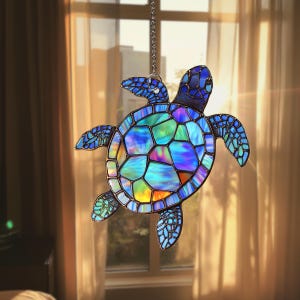 Könnte beinhalten: Ein Meeresschildkröten-Sonnenfänger aus Buntglas mit irisierenden blauen, grünen und violetten Glaspaneelen, die durch dunkle Metalllötstellen verbunden sind. Die Schildkröte hängt an einer silberfarbenen Metallkette und zeigt ein Mosaikmuster.