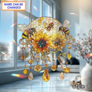 May include: A decorative suncatcher featuring sunflowers, bees, and honeycombs. The design includes the text "Laura" and "Style 1". The suncatcher is adorned with hanging crystal-like accents and is suspended by a chain.