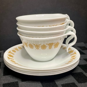 May include: A set of four white ceramic teacups and saucers with a gold floral pattern. The cups have a rounded shape and a handle on the side. The saucers have a scalloped edge.