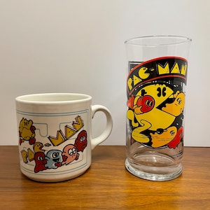 May include: A white ceramic mug and a clear glass with Pac-Man graphics. The mug has a yellow Pac-Man and four ghosts in a maze. The glass has a yellow Pac-Man with a ghost in the background.
