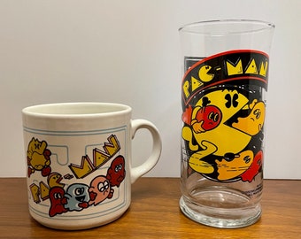 May include: A white ceramic mug and a clear glass with Pac-Man graphics. The mug has a yellow Pac-Man and four ghosts in a maze. The glass has a yellow Pac-Man with a ghost in the background.