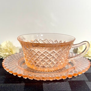 May include: A pink glass teacup and saucer set with a diamond pattern. The cup and saucer are both made of clear glass with a pink tint.