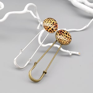 Könnte beinhalten: Zwei Sicherheitsnadeln mit runden Leopardenmuster-Designs. Eine Nadel hat eine silberne Oberfläche, die andere eine bronzefarbene. Die Nadeln sind vor einem grauen Hintergrund mit weißen Ästen platziert.