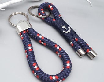 May include: Two nautical-themed keychains. One features a loop of navy blue, red, and white rope with a silver-toned metal cap. The other has a navy blue band with a silver anchor emblem and rope ends. Both have silver key rings.