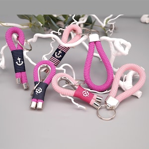 Könnte beinhalten: Eine Sammlung von fünf Seil-Schlüsselanhängern in Pink- und Magentatönen. Jeder Schlüsselanhänger besteht aus einer geflochtenen Kordelschlaufe, einem Metallring und einem dunkelblauen oder pinken Stoffband mit silbernem Anker- oder Kleeblatt-Symbol.