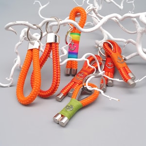 Könnte beinhalten: Eine Sammlung von orangefarbenen Seil-Schlüsselanhängern mit silberfarbenen Akzenten. Einige Schlüsselanhänger haben einen Regenbogenstreifen, ein grünes Band oder einen Ankeranhänger. Die Schlüsselanhänger sind vor einem grauen Hintergrund mit weißen Ästen angeordnet.