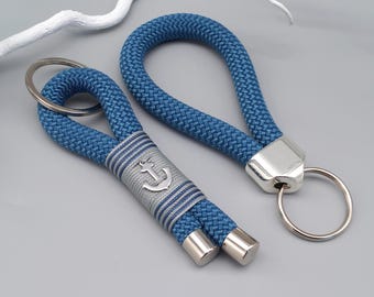 May include: Two blue rope keychains with silver accents. One keychain features a silver anchor emblem on a gray and blue striped band. Both keychains have silver key rings and end caps. The keychains are on a gray background.