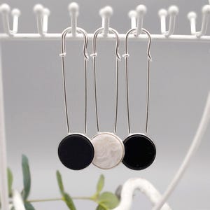 Könnte beinhalten: Drei silberfarbene Sicherheitsnadel-Ohrringe. Zwei Ohrringe haben schwarze runde Anhänger, und einer hat einen weißen, marmorierten Anhänger. Die Ohrringe hängen an einem weißen Gestell, mit grünem Laub im Hintergrund.