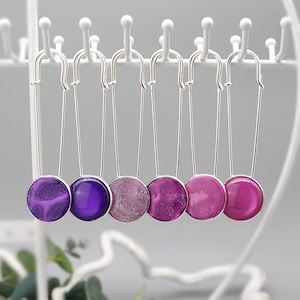 Könnte beinhalten: Sieben silberfarbene Sicherheitsnadeln, jede mit einem kreisförmigen Charm in Lila- und Rosatönen. Die Farben reichen von tiefem Violett bis zu hellem Magenta. Die Nadeln werden auf einem weißen Gestell präsentiert. Die Charms sind aus Kunstharz.