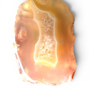 May include: A translucent, polished agate slice featuring warm amber, peach, and cream tones. The stone displays natural concentric banding and a central crystalline geode formation that allows light to pass through its smooth, irregular surface.