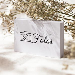 May include: A rectangular white card featuring a black line-art icon of a camera with a heart in the centre, followed by the black cursive text "Hochzeits-Fotos". The card is positioned upright with a stack of identical cards visible behind it.