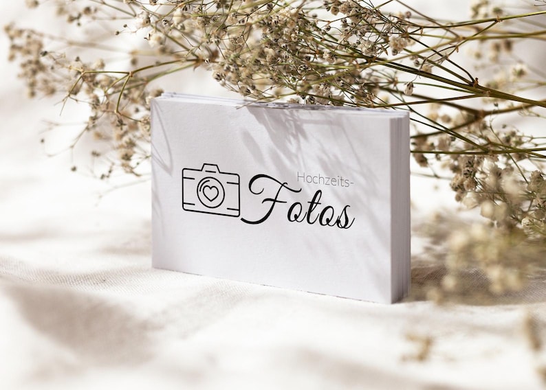 May include: A rectangular white card featuring a black line-art icon of a camera with a heart in the centre, followed by the black cursive text "Hochzeits-Fotos". The card is positioned upright with a stack of identical cards visible behind it.
