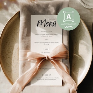 May include: A white menu card with black text titled Menü, placed on a beige linen napkin tied with a light pink silk ribbon. The card rests on a speckled ceramic plate flanked by a silver fork on the left and a knife and spoon on the right.