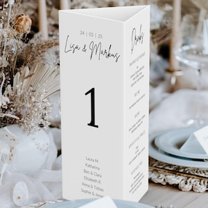 May include: A white, triangular table number card with the number 1 in bold black font. The card features the names "Lisa & Markus" and a date. Additional text includes a list of names and a drinks menu.