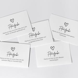May include: Five white rectangular cards with black text and a hand-drawn heart icon. Each card features the heading Fotoaufgabe followed by specific German instructions for taking photos, including tasks like photographing a guest who married recently.