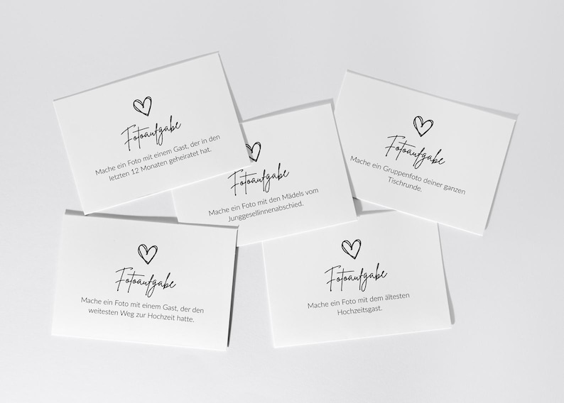 May include: Five white rectangular cards with black text and a hand-drawn heart icon. Each card features the heading Fotoaufgabe followed by specific German instructions for taking photos, including tasks like photographing a guest who married recently.