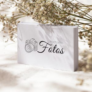 May include: A stack of white rectangular business cards featuring a black line drawing of a camera and the text Hochzeits-Fotos printed in a combination of sans-serif and cursive fonts, positioned against a background of dried white flowers.
