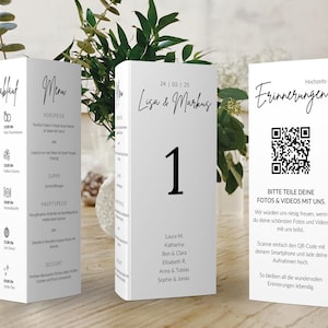May include: White wedding table cards with black text. One card displays the wedding schedule, another the menu, and a third card has a QR code and text. The center card has the number 1 and names. The cards are on a wooden surface.