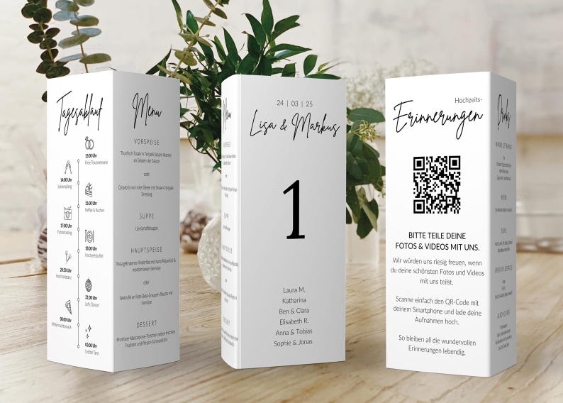 May include: White wedding table cards with black text. One card displays the wedding schedule, another the menu, and a third card has a QR code and text. The center card has the number 1 and names. The cards are on a wooden surface.