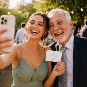 May include: A small white rectangular card featuring a hand-drawn heart icon above the text 'Fototaufgabe' and additional printed instructions, held by two individuals posing for a selfie with a smartphone at an outdoor event.