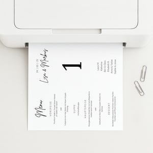 May include: A white printer is printing a menu card. The card has the names "Lisa & Markus" and the number "1" in black. Two silver paper clips are on the white surface.