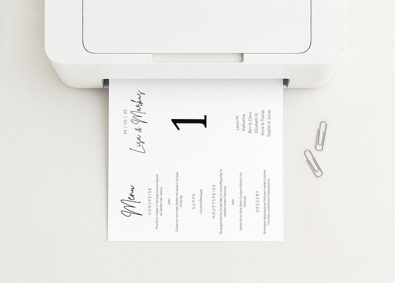 May include: A white printer is printing a menu card. The card has the names "Lisa & Markus" and the number "1" in black. Two silver paper clips are on the white surface.