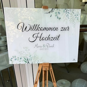 Könnte beinhalten: Ein weißes Schild auf einer Holzstaffelei mit dem Schriftzug "Willkommen zur Hochzeit" in eleganter Schrift. Das Schild enthält auch die Namen "Maria & Daniel" und das Datum "09.07.2022" mit einem floralen Design.