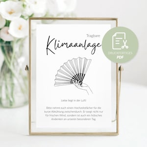 May include: A framed print with the words "Tragbare Klimaanlage" and a drawing of a hand holding a fan. The print is in a gold frame, with a vase of white flowers in the background.