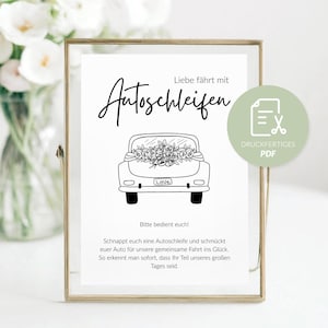 May include: A framed print with black text and a car illustration. The text reads "Liebe fährt mit Autoschleifen" and "Bitte bedient euch!" A car illustration is in the center, with a bouquet of flowers in the back. A PDF icon is in the upper right.