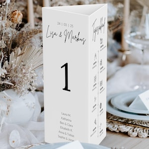 May include: A white, triangular table number card with the number 1 in bold black font. The card features the names "Lisa & Markus" and the date "24/03/25" in black script. Additional text and icons are printed on the sides. Wedding decor.