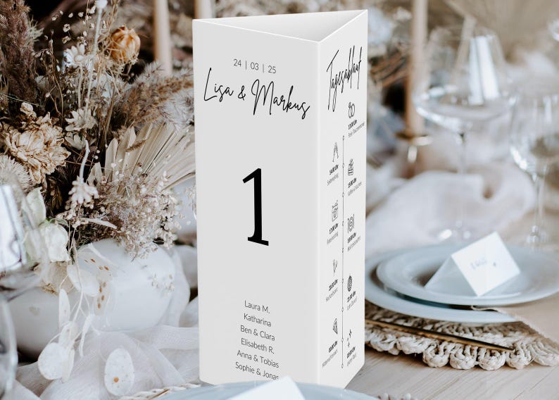 May include: A white, triangular table number card with the number 1 in bold black font. The card features the names "Lisa & Markus" and the date "24/03/25" in black script. Additional text and icons are printed on the sides. Wedding decor.