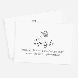 May include: Two white cards with a black line drawing of a camera and the word "Fotoaufgabe" in script. The text below reads, "Mache ein Foto mit einem Gast, der in den letzten 12 Monaten geheiratet hat."