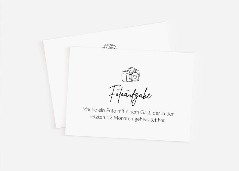 May include: Two white cards with a black line drawing of a camera and the word "Fotoaufgabe" in script. The text below reads, "Mache ein Foto mit einem Gast, der in den letzten 12 Monaten geheiratet hat."