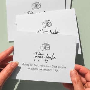 May include: White cards with a camera illustration and the text "Fotoaufgabe" in a handwritten font. The cards are held by hands against a light green background. The text on the card reads "Mache ein Foto mit einem Gast, der ein originelles Accessoire tr&auml;gt."