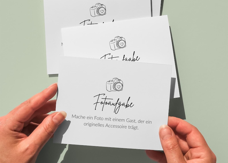 May include: White cards with a camera illustration and the text "Fotoaufgabe" in a handwritten font. The cards are held by hands against a light green background. The text on the card reads "Mache ein Foto mit einem Gast, der ein originelles Accessoire tr&auml;gt."