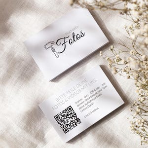 May include: Two white rectangular cards with black text and graphics. The top card features a camera icon and the text "Hochzeits-Fotos". The bottom card displays a QR code and the text "BITTE TEILE DEINE FOTOS & VIDEOS MIT UNS. Scanne den QR-Code mit deinem Smartphone und lade deine Aufnahmen hoch. Lieben Dank! Lisa & Markus".