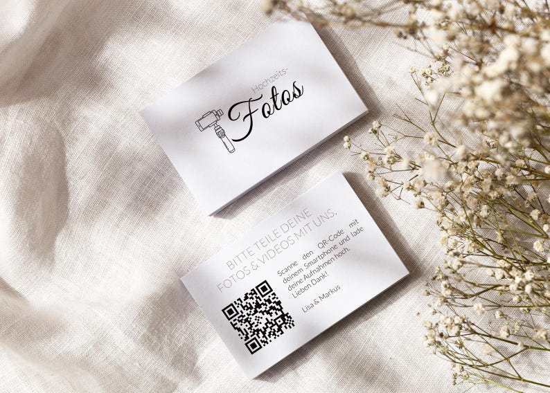 May include: Two white rectangular cards with black text and graphics. The top card features a camera icon and the text "Hochzeits-Fotos". The bottom card displays a QR code and the text "BITTE TEILE DEINE FOTOS & VIDEOS MIT UNS. Scanne den QR-Code mit deinem Smartphone und lade deine Aufnahmen hoch. Lieben Dank! Lisa & Markus".