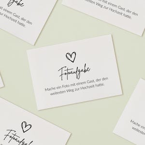 May include: Multiple white rectangular cards featuring black text and a hand-drawn heart icon. The text reads "Fotoaufgabe" followed by "Mache ein Foto mit einem Gast, der den weitesten Weg zur Hochzeit hatte." The cards are arranged in a scattered pattern.