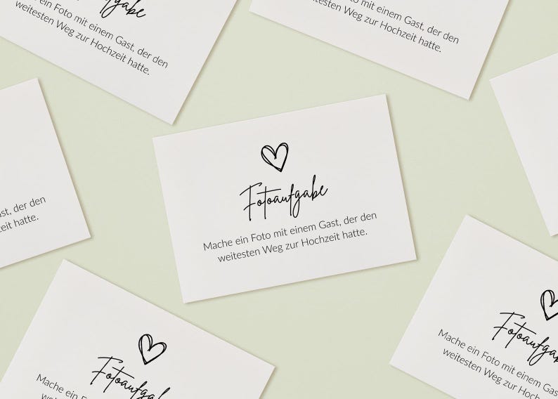 May include: Multiple white rectangular cards featuring black text and a hand-drawn heart icon. The text reads "Fotoaufgabe" followed by "Mache ein Foto mit einem Gast, der den weitesten Weg zur Hochzeit hatte." The cards are arranged in a scattered pattern.