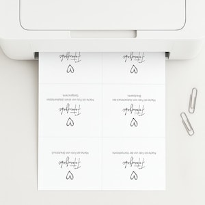 May include: A white printer feeding a sheet of paper with four identical cards featuring the German text Fototaufgabe and a hand-drawn heart icon. Two silver metal paper clips are positioned to the right of the printed paper sheet.