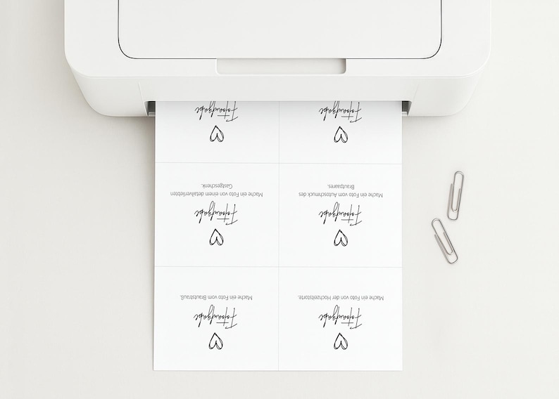 May include: A white printer feeding a sheet of paper with four identical cards featuring the German text Fototaufgabe and a hand-drawn heart icon. Two silver metal paper clips are positioned to the right of the printed paper sheet.