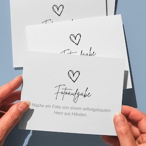 May include: A stack of white rectangular cards featuring a black hand-drawn heart icon above the text "Fotoaufgabe" and the German sentence "Mache ein Foto von einem selbstgebauten Herz aus H&auml;nden." The cards are held against a solid light blue background.