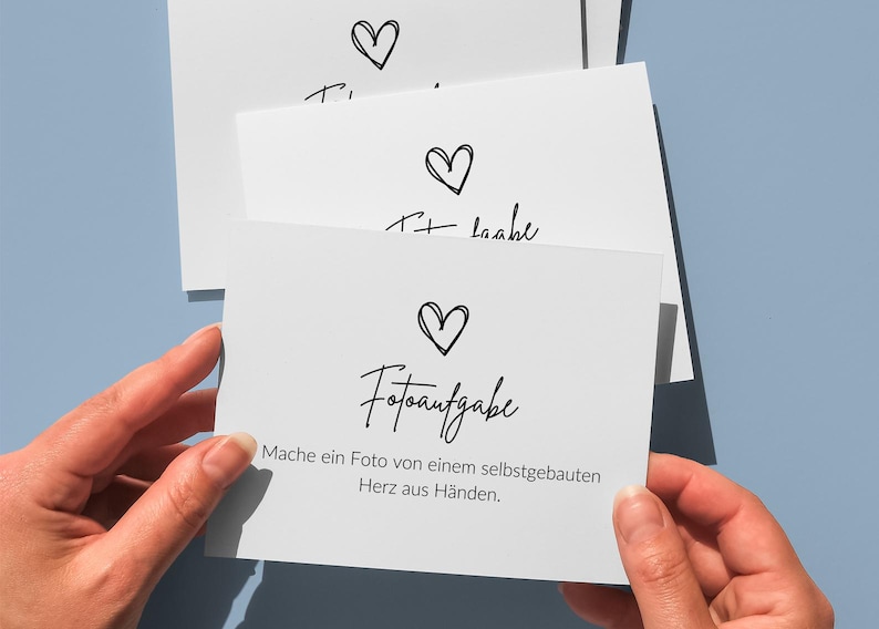 May include: A stack of white rectangular cards featuring a black hand-drawn heart icon above the text "Fotoaufgabe" and the German sentence "Mache ein Foto von einem selbstgebauten Herz aus H&auml;nden." The cards are held against a solid light blue background.